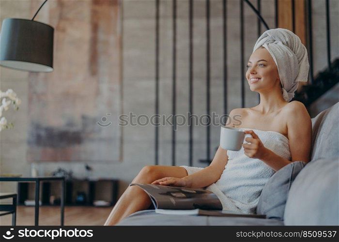 Photo of relaxed smiling woman enjoys drinking tea, reads magazine and poses on comfortable sofa in living room, enjoys spare time during day off, wears minimal makeup, has well cared healthy skin