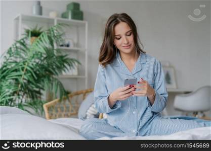 Photo of relaxed young European lady in pyjamas enjoys audio playlist, listens music romantic songs in earphones, uses modern cell phone, sits on comfortable bed against domestic atmosphere.