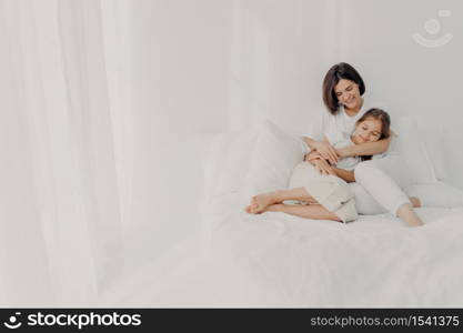 Photo of relaxed young mother embraces daughter with love, pose in pyjamas bare foot, have pleasant smiles, being in spacious white bedroom, have good relationships, wake up late in morning.