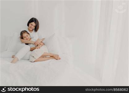 Photo of relaxed young mother embraces daughter with love, pose in pyjamas bare foot, have pleasant smiles, being in spacious white bedroom, have good relationships, wake up late in morning.