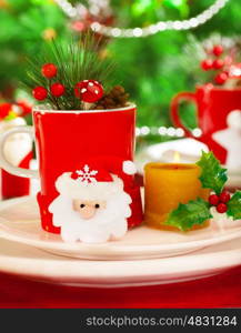 Photo of romantic wintertime table setting, red cup for tea full of different Christmastime decoration, small Santa Clause decor with yellow candle on white festive plate on Christmas tree background