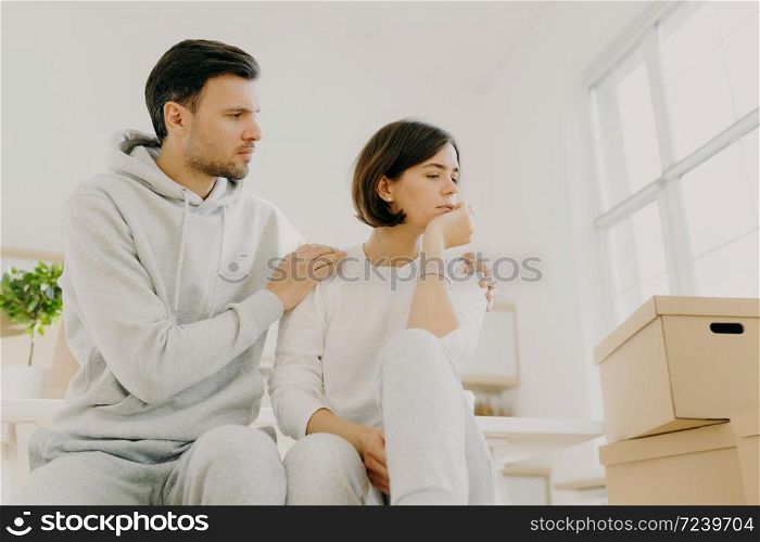 Photo of sad woman complains about problems, man calms, embraces wife, tired after bringing many boxes with things to their new home, wear casual clothes, being stressful. Family problems concept