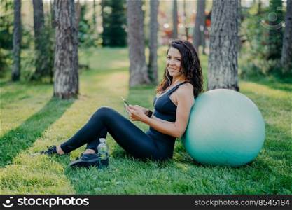Photo of satisfied brunette young woman sits back to big fitness ball, uses mobile phone for listening music, poses on green grass in forest during sunny day, wears sports clothes. Sport concept