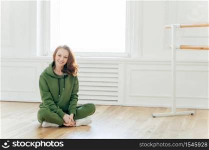 Photo of satisfied foxy European woman keeps legs crossed, poses near ballet barre, takes rest after dancing, wears green tracksuit, white sportshoes, stretches herself, models in dancing studio