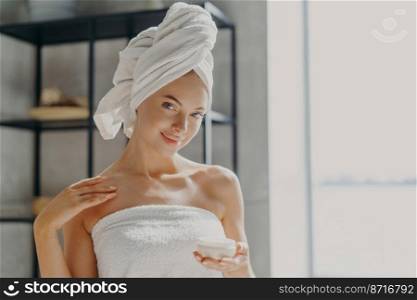 Photo of satisfied young woman has healthy pure skin, applies body cream, uses skin treatment to moisturize, stands wrapped in towel, smiles gently. Personal care, beauty and wellness concept