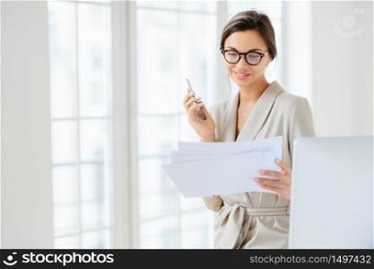 Photo of satisfied young woman with dark hair dressed in business suit, focused in papers, works in office, holds modern cellphone, wears optical glasses for good vision, has pleased expression