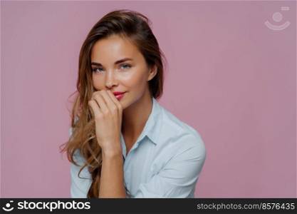 Photo of self assured attractive woman with long hair, keeps hand on lips, dressed in blue shirt, looks at camera with blue eyes, wears makeup, poses against purple background. Human face expressions