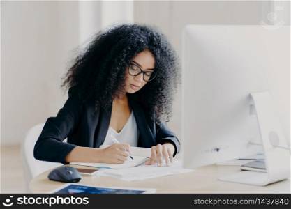 Photo of serious Afro woman writes in papers, sits at table with modern computer, creats article in newspaper, wears glasses and eyewear, poses indoor, works remotely. People and job concept