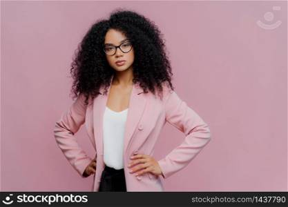 Photo of serious curly haired woman looks confidently at camera, wears formal neat clothes, keeps both hands on waist, wears optical glasses, poses over purple background. People, style concept