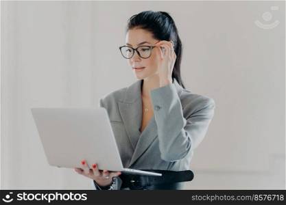 Photo of serious executive worker concentrated in laptop screen, wears transparent glasses for vision correction, browses internet web page, dressed in elegant formal clothes stands against white wall