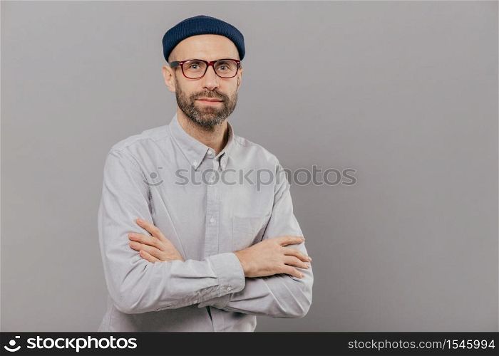 Photo of serious male keeps arms folded over chest, dressed in white formal shirt, fashionable hat, poses for making photography over grey wall with free space for your advertisement or promotion