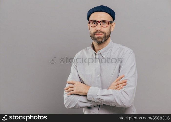Photo of serious male keeps arms folded over chest, dressed in white formal shirt, fashionable hat, poses for making photography over grey wall with free space for your advertisement or promotion