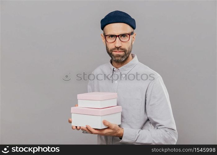 Photo of serious male model dressed in festive clothes, holds gifts in hands, going to congratulate friend with birthday, ready for party wears spectacles isolated over grey wall. This present for you