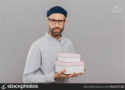 Photo of serious male model dressed in festive clothes, holds gifts in hands, going to congratulate friend with birthday, ready for party wears spectacles isolated over grey wall. This present for you