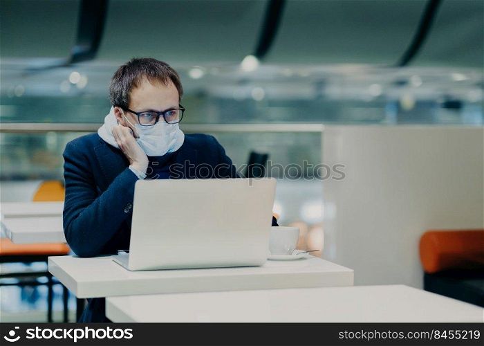 Photo of serious man concentrated in screen of laptop computer, works from distance during coronavirus outbreak, wears protective mask not to spread disease, drinks coffee, sits at white table