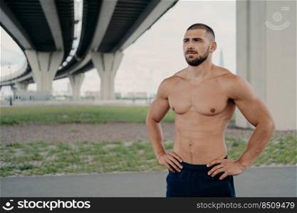 Photo of shirtless handsome bearded man with naked torso poses outdoor keeps hands on waist concentrated into distance goes in for sport, has morning workout, proud to be strong, has perfect abs