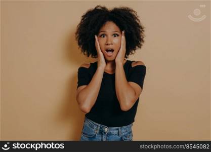 Photo of shocked pretty dark skinned curly haired lady holding hands on cheekbones not believe becouse of discount low prices, wear trendy black tshirt isolated over dark beige studio background. Photo of shocked pretty dark skinned curly haired lady holding hands on cheekbones