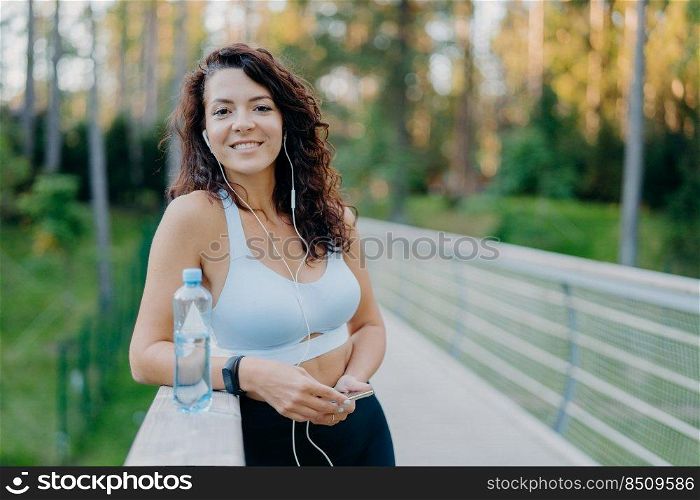 Photo of slim beautiful curly woman dressed in cropped top and leggings enjoys listening music holds mobile phone drinks fresh water from bottle poses at bridge poses against nature background