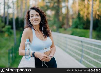 Photo of slim beautiful curly woman dressed in cropped top and leggings enjoys listening music holds mobile phone drinks fresh water from bottle poses at bridge poses against nature background