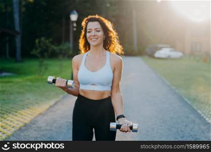 Photo of slim brunette young woman raises dumbbells has morning workout poses against sunrise dressed in cropped top and leggings works on arms muscles smiles positively leads sporty lifestyle
