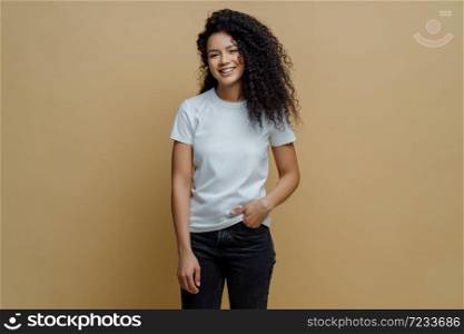 Photo of slim cheerful African American woman with curly hair, smiles happily, being in good mood, wears white t shirt and jeans, keeps hand in pocket, has slim figure, isolated on beige background