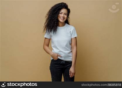 Photo of slim cheerful African American woman with curly hair, smiles happily, being in good mood, wears white t shirt and jeans, keeps hand in pocket, has slim figure, isolated on beige background