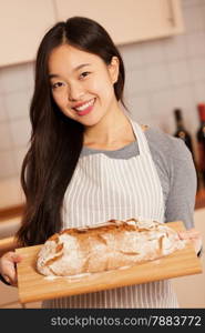 photo of smiling asian woman holding fresh baked bread on a wooden tray at home