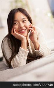 photo of smiling asian woman speaking at phone while lying on the sofa at home