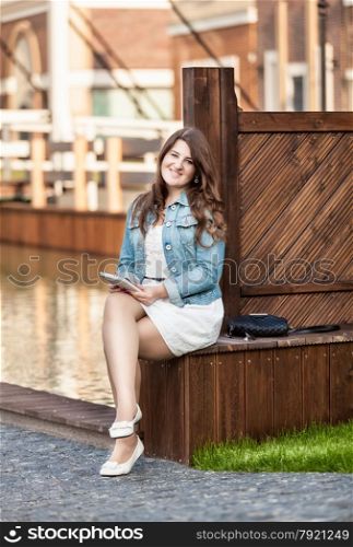 Photo of smiling brunette woman sitting on bridge and using tablet