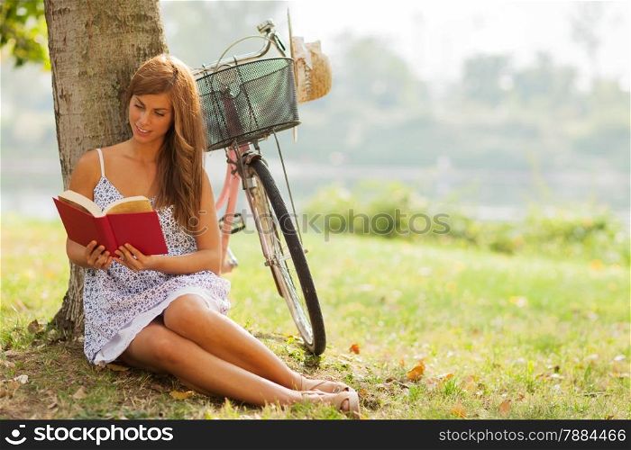 photo of smiling caucasian woman reading a book next to a tree where her bike is standing