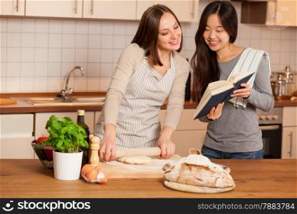 photo of smiling caucasian woman working on a dough while her friend is reading a cookbook