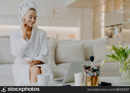 Photo of smiling woman applies nourishing cream on face looks in laptop screen gives advice of skin care dressed in bathrobe and towel sits at comfortable sofa has rejuvenation procedure. Cosmetology