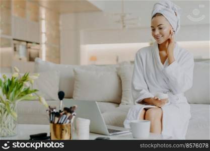 Photo of smiling woman applies nourishing cream on face looks in laptop screen gives advice of skin care dressed in bathrobe and towel sits at comfortable sofa has rejuvenation procedure. Cosmetology