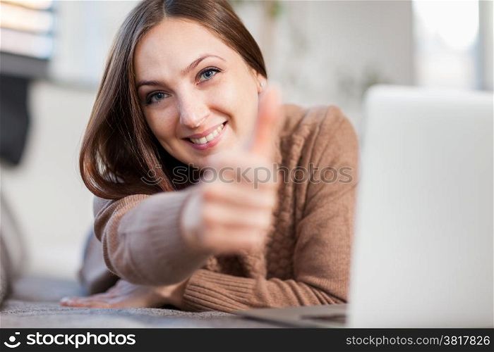 photo of smiling woman doing positive thumb gesture while lying on a couch and chatting with computer