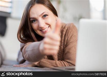 photo of smiling woman doing positive thumb gesture while lying on a couch and chatting with computer