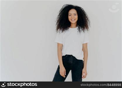 Photo of smiling woman with curly dark hair, has slim figure, wears white t shirt and black jeans, has glad face expression, models against white background, copy space for your advertising content