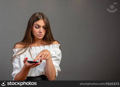 Photo of smiling woman with long dark hair holding and using black mobile phone isolated over gray background