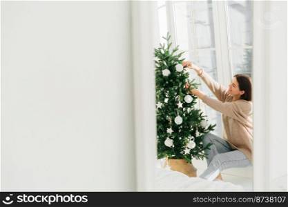 Photo of smiling young female decorates Christmas tree at home, poses against big window, white copy space on left side. Celebration, winter time and people concept. Festive mood, cozy interior