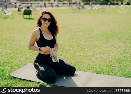 Photo of sporty Caucasian woman sits on mat drinks water after exercising feels refreshed and satisfied goes in for sport outdoor during summer day wears sunglasses and activewear. Healthy lifestyle