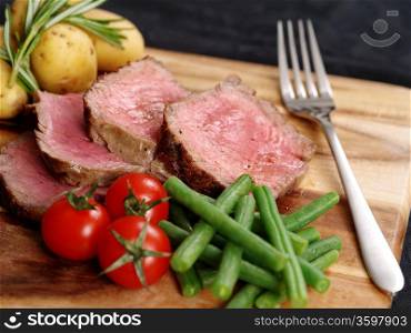 Photo of steak dinner with thick slices of sirloin, cherry tomatoes, green beans and potatoes on a wooden board.