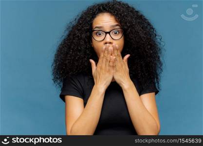 Photo of stupefied young female covers mouth, has bated breath, bugged eyes, Afro hair, cannot believe in shocking news, wears casual black t shirt, stands over blue background. Omg concept.