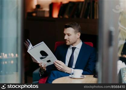 Photo of successful businessman recreats at coffee shop, drinks aromatic hot beverage, focused into magazine, waits for other order, dressed in formal clothes, has stubble and appealing look