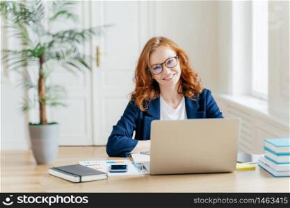 Photo of successful businesswoman with red hair, wears optical glasses for vision correction, sits at desktop in front of opened laptop, thinks on organization plan, surrounded with paper documents