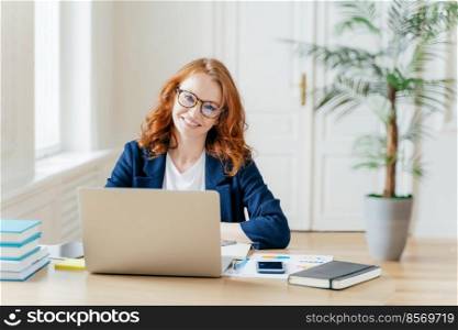 Photo of successful businesswoman with red hair, wears optical glasses for vision correction, sits at desktop in front of opened laptop, thinks on organization plan, surrounded with paper documents