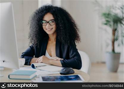 Photo of successful female manager in elegant suit, smiles pleasantly, makes notes, sits at desktop, drinks coffee, poses at computer, smiles positively, poses in office interior. People and work