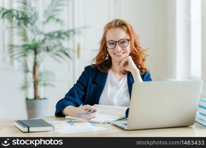 Photo of successful ginger female freelancer has remote work, watches webinar online on laptop computer, writes down information in notepad, smiles happily poses at workplace dressed in formal clothes