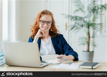 Photo of successful ginger female freelancer has remote work, watches webinar online on laptop computer, writes down information in notepad, smiles happily poses at workplace dressed in formal clothes