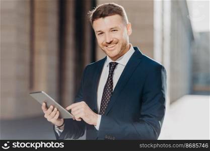 Photo of successful male banker does remote work on digital tablet, has happy expression, dressed in formal black suit, has happy look at camera. Positive young man makes transactions online