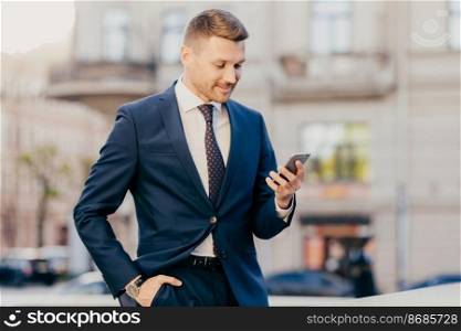 Photo of successful male entrepreneur holds modern smart phone in hands, reads news from financial exchange, browses internet, makes online payments, has work break, going to cafe or restaurant
