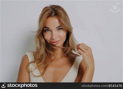 Photo of tender shy woman holds hair wears minimal makeup dressed in casual wear has tender expression dressed casually poses against grey background. Women beauty and face expressions concept. Photo of tender shy woman holds hair wears minimal makeup dressed in casual wear